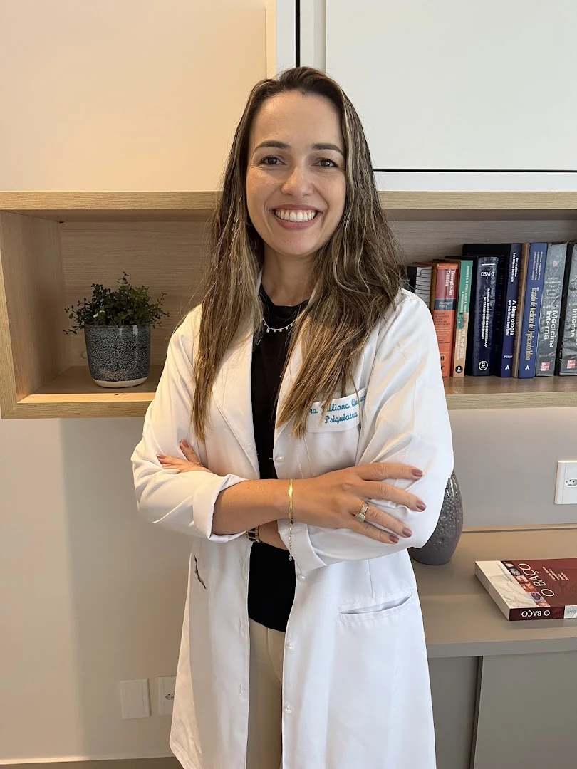 Dra. Julliana Quintas, psiquiatra, sorrindo em seu consultório em Icaraí, de jaleco, diante de estante com livros de medicina